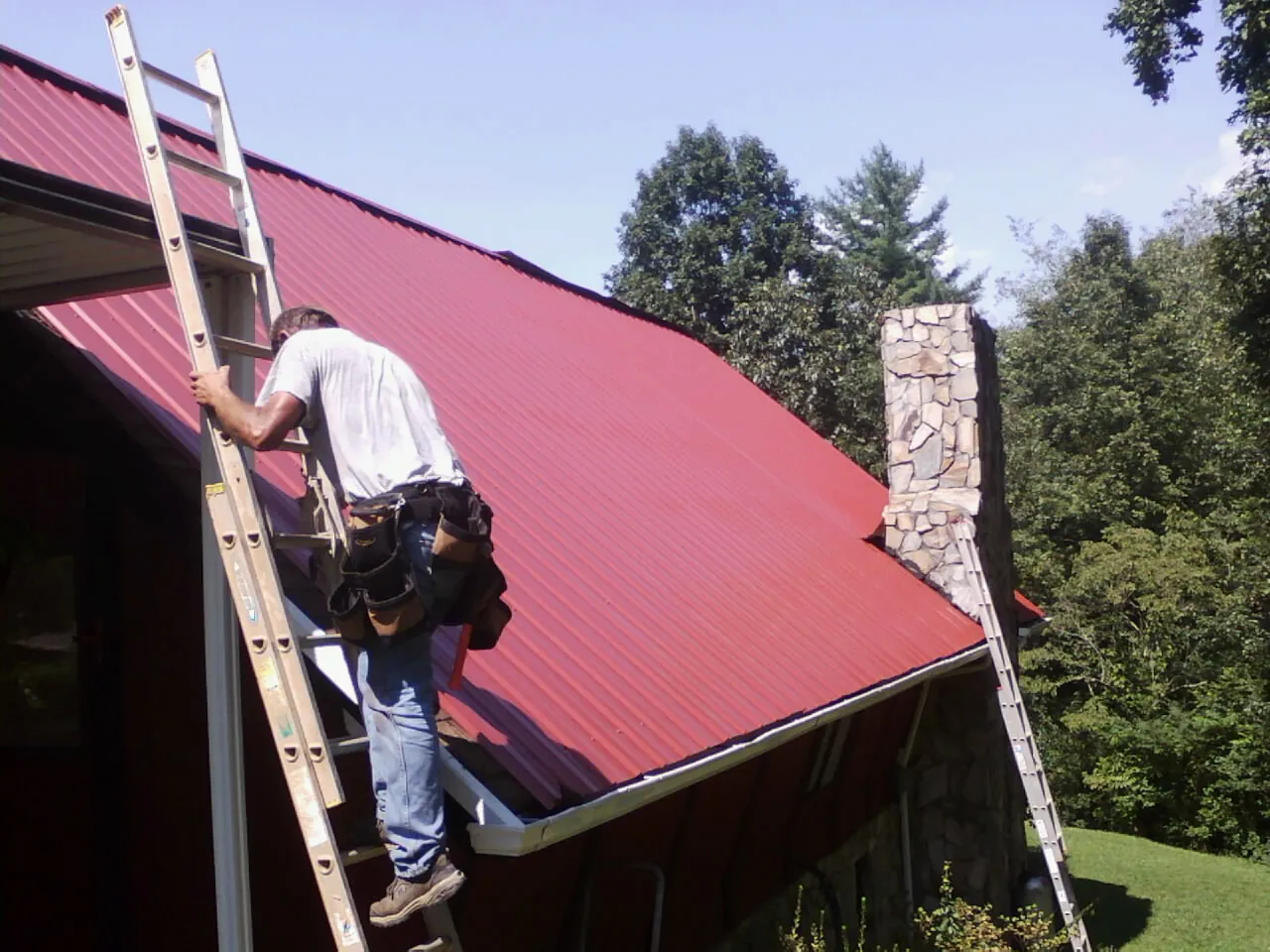 Roofer on a ladder beside a red metal roof and stone chimney — Soffit & Fascia Repair by Roofing Brothers in Canmer